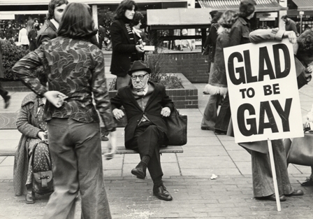 High street scene with shoppers and protestors, including a prominent Glad to Be Gay banner