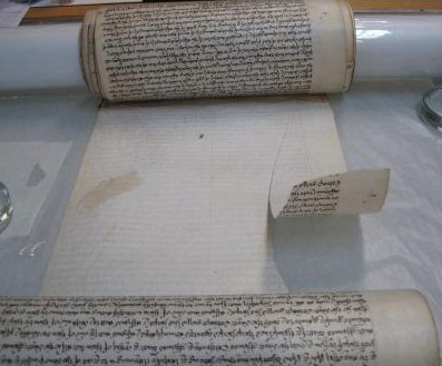 Image of a large parchment roll, lying on its front with a long tear running parallel to right hand side. The roll bears densely written text in a brown ink.