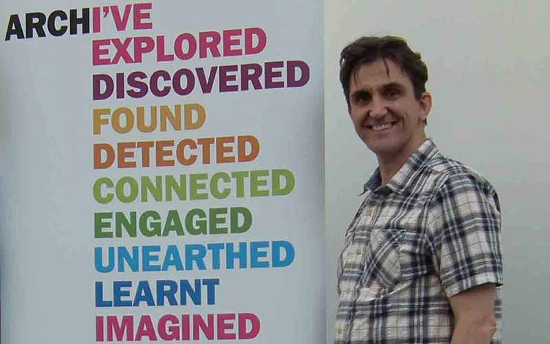 Stephen McGann stands in front of and Explore Your Archive poster