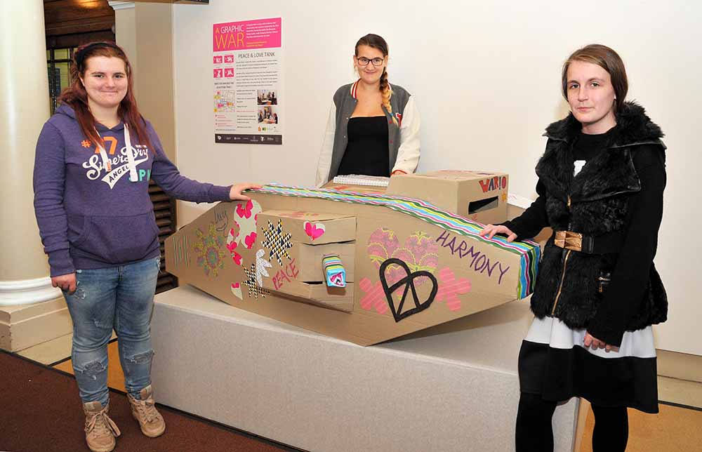 Three young women stand next to a decorated cardboard tank