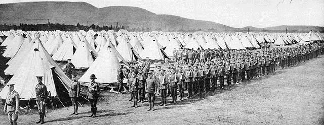 A black-and-white photo showing a field of white tents as far as the eye can see, and a long line of soldiers conducting military exercises in the foreground.