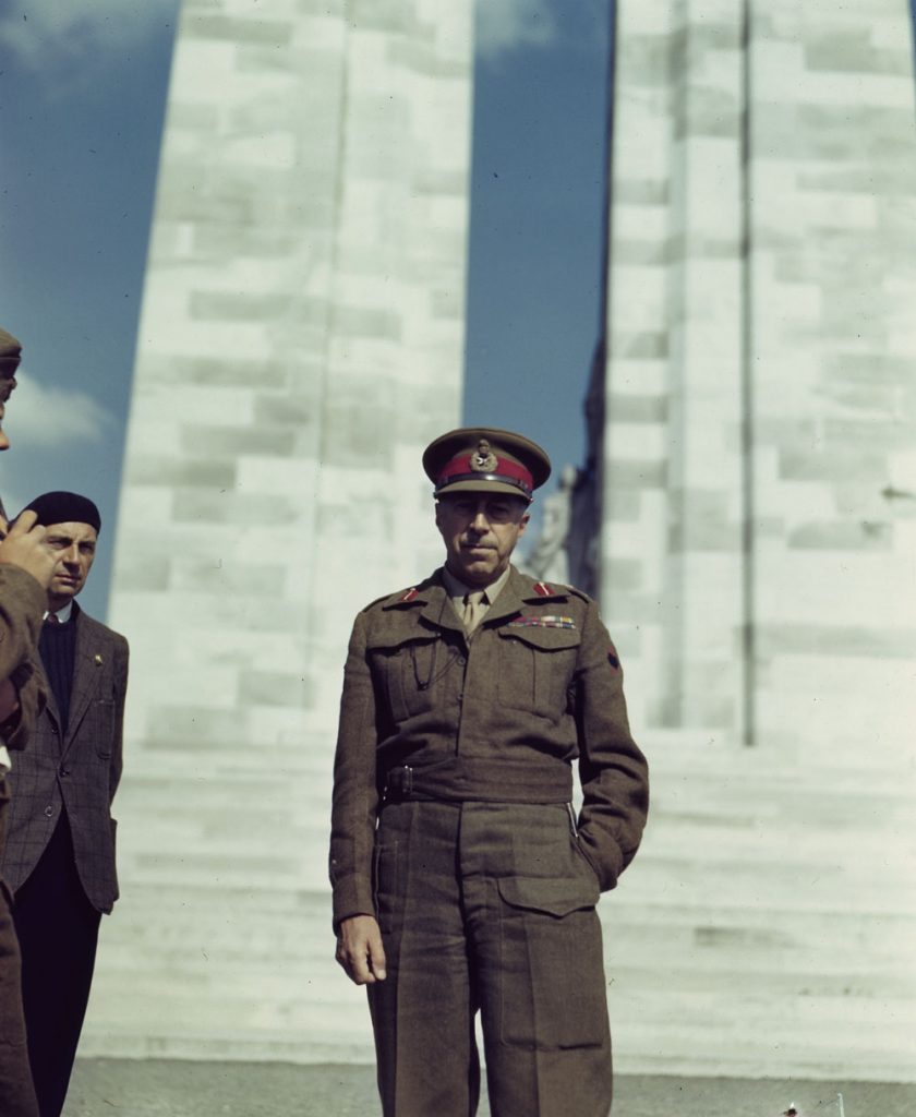 A colour photograph of man standing in front of a large stone structure. Two people are on the left side of the photograph, one is in uniform and mostly cut off and the other is wearing a vest, sweater and beret.