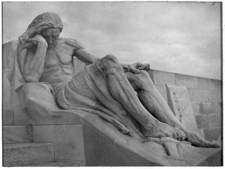 A black-and-white photograph of a dramatic view of a larger-than-life sculpture from the Vimy Memorial, a man in mourning with his foot resting on a sword. In the background are side panels bearing the names of Canadian dead.