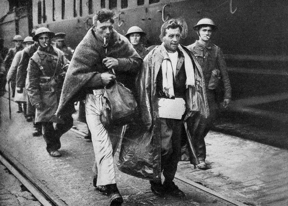 Black and white photograph of a group of men walking alongside a train; one is wrapped in a blanket and is smoking a pipe