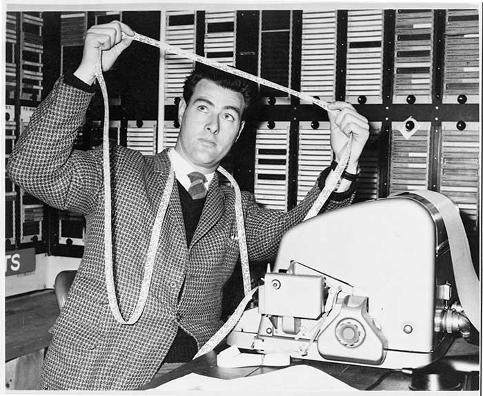 1960s image of a man examining digital tape emerging from a machine