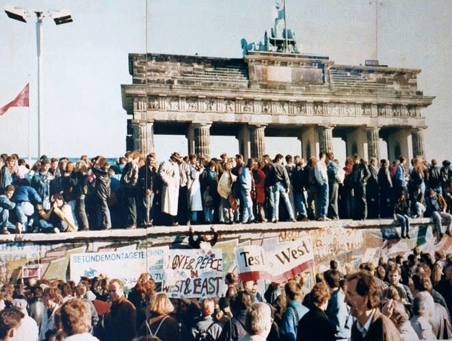 West and East Berliners are shown standing on top of the Berlin Wall. The Brandenburg Wall is shown behind the wall. in the foreground a peaceful crowd of people are milling around. Some are holding banners - one reads:' Love and Peace in West and East'
