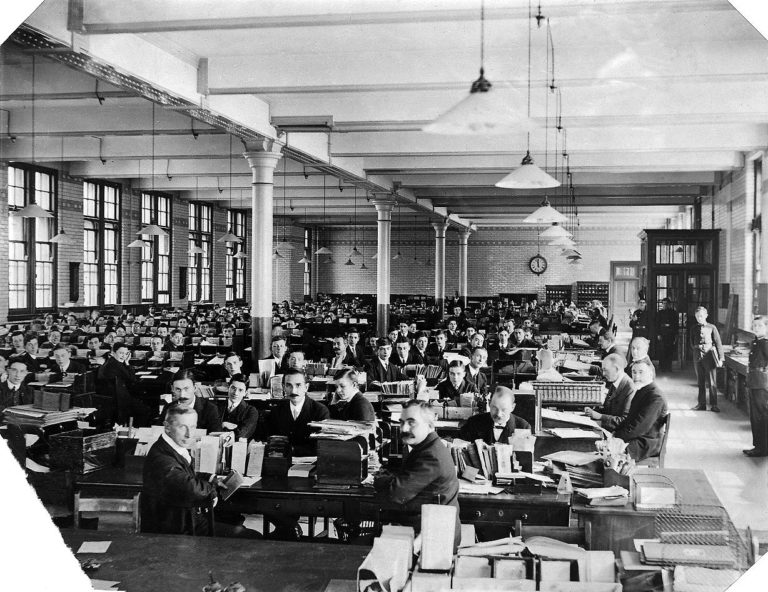 Photograph of staff at the Post Office Savings Bank, Blythe Road, Kensington, London dated 1910 to 1920. Shows around 150 men sitting at rows of desks surrounded by paperwork. Document reference NSC 27/2/12.
