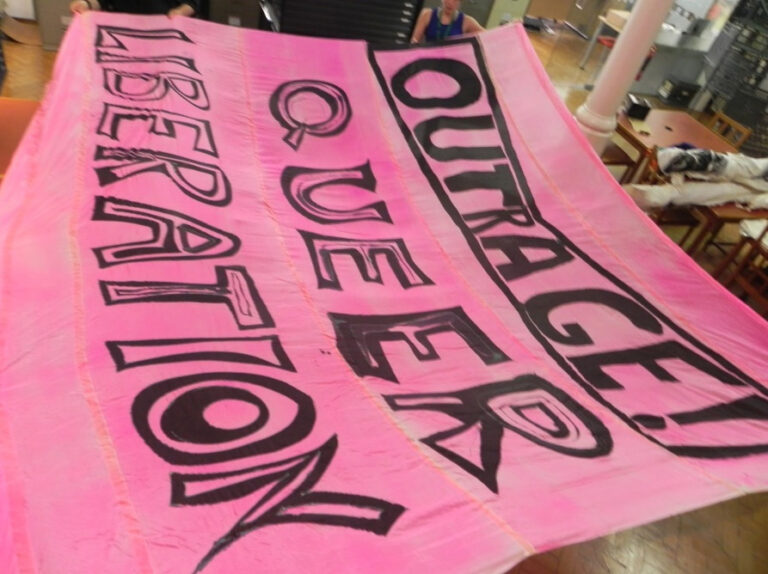 A pink banner with black lettering that says ‘Outrage! Queer Liberation’.