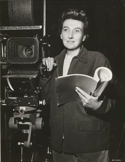 Muriel Box on set standing against a camera and holding a script.