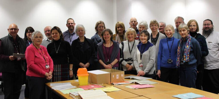 A group of volunteers, consisting of 20 men and women and staff from The National Archives, standing around a table upon which are spread out documents from the archives.