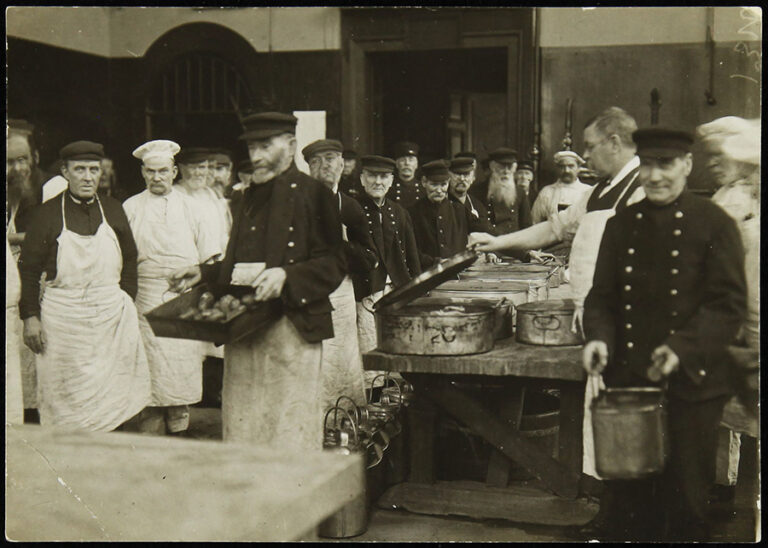 A group of Chelsea Pensioners dressed in uniform queue up in a dining room to be served their food rations.