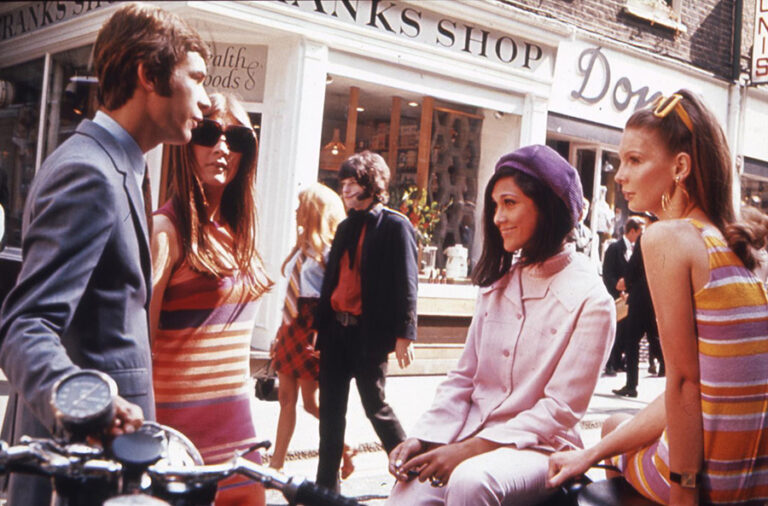 A group of fashionable people sit talking on a 1960s London street.