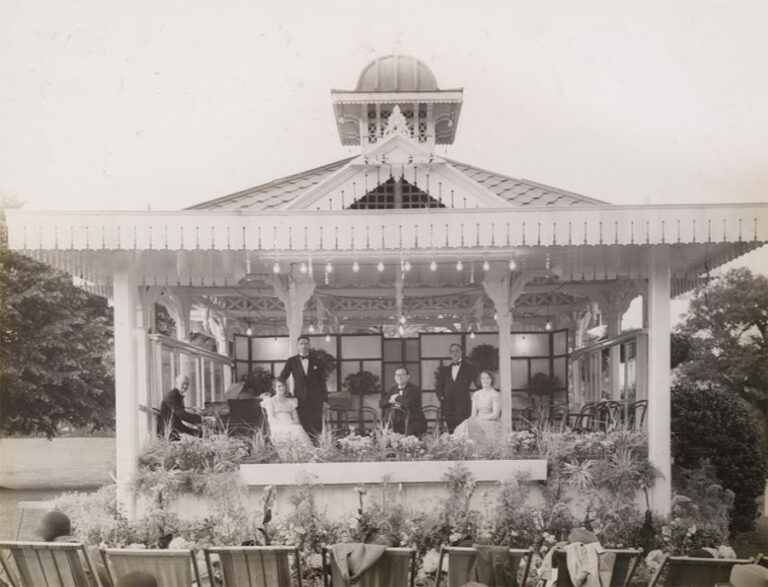A group of smartly dressed musicians pose for a photograph in a pavilion, in front of a small crowd in deckchairs.