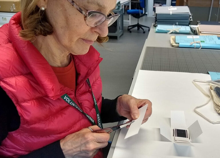 A middle-aged woman wearing a volunteer lanyard is shown cutting a piece of card.