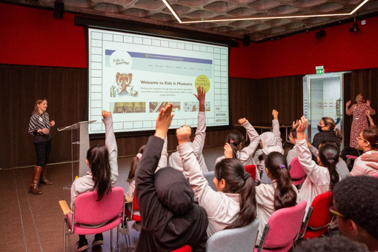 In a lecture hall a group of school children interact with a teacher who is standing in front of a presentation on a big screen. The children have raised hands.