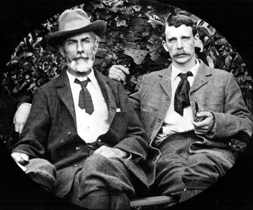 Posed photographic portrait of two middle-aged men who are sitting on a bench in a garden.