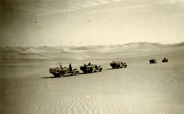 A sepia tinted photograph showing five army patrol vehicles travelling in line through the desert.