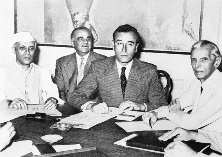 Politicians Jawaharlal Nehru, Muhammad Ali Jinnah, Lord Mountbatten are seated together at a table as Mountbatten signs a document.