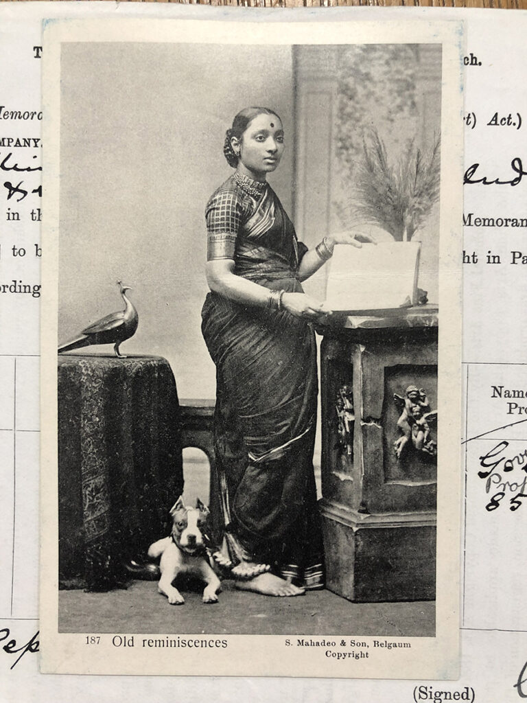 A woman wearing a sari stands in front of an open ledger. She is barefoot and a dog sits at her feet.