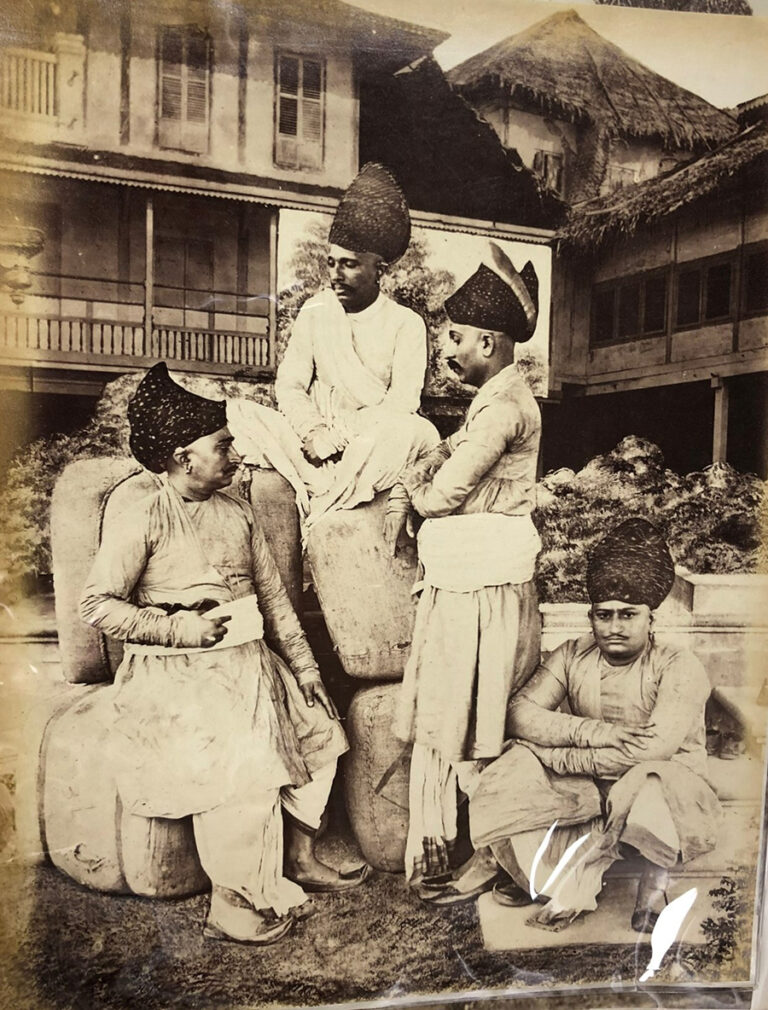 Four men sitting on bales in front of a house.