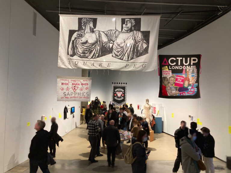 People in a gallery stand in groups talking and looking at artwork on walls and banners hung from the ceiling.