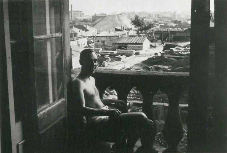 John Vassall sits shirtless on a balcony of a property overlooking other small buildings.