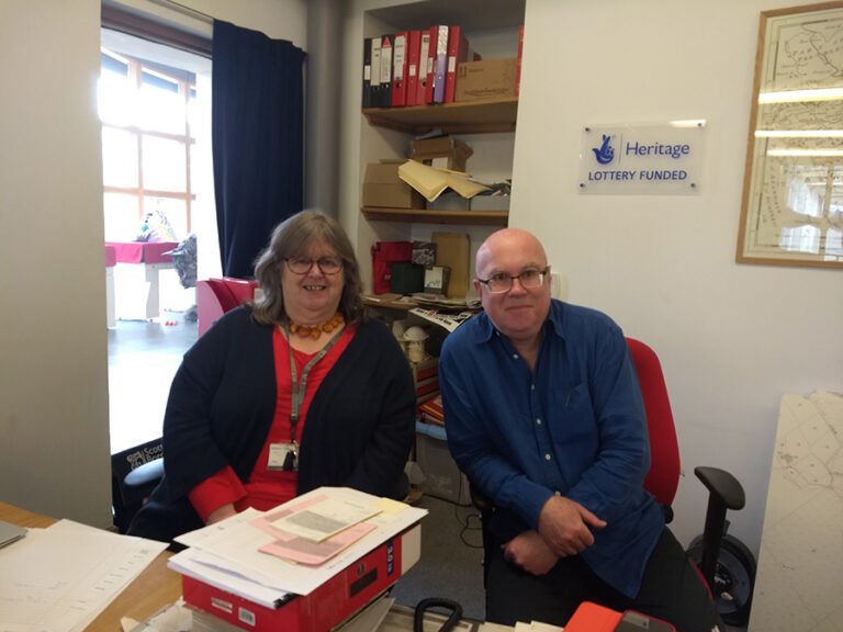 Two middle-aged people sit side by side in a small office.