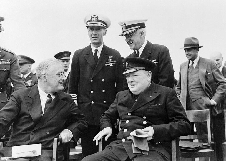 President Roosevelt and Winston Churchill are shown in conversation with a group of men around them in military uniform.