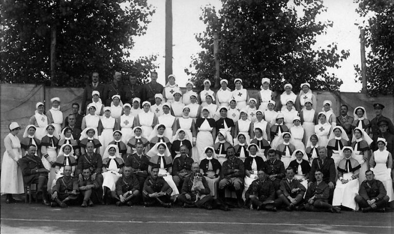 The nurses and staff of Queen Alexandra’s Imperial Military Nursing Service stand for a posed group photograph. They are arranged over five rows, with the front row seated.