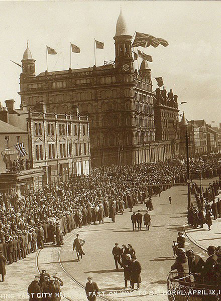 Crowds line the streets in front of a tall building displaying a Union Jack flag.