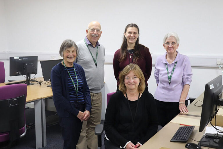 Four women and one man stand among desks and computers smiling for the camera.