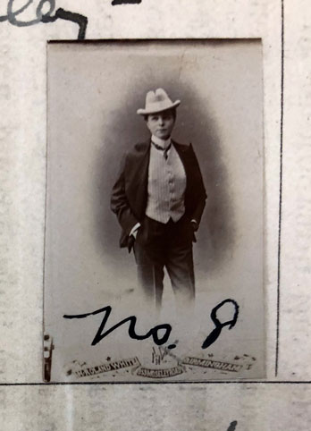 A woman poses with her hands in her pockets while wearing a suit and a white hat.