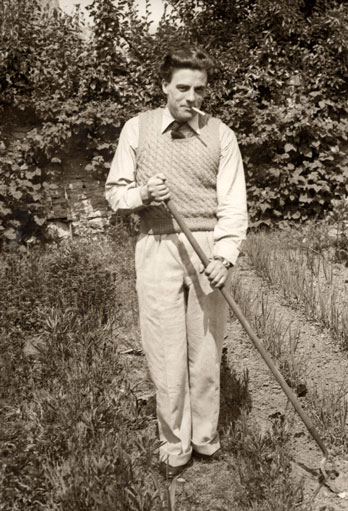 A man with a cigarette in his mouth stands in a ploughed field, holding a rake.