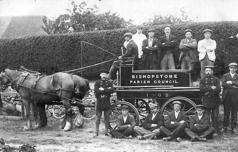 Black and white photograph of a horse-drawn fire engine. A group of men stand on and around the fire engine, posing with their arms crossed for the camera. Two of the men are in uniform