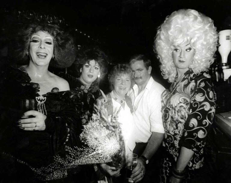Three stylishly-dressed drag queens, one smiling and one smirking, stand beside a man and a woman.