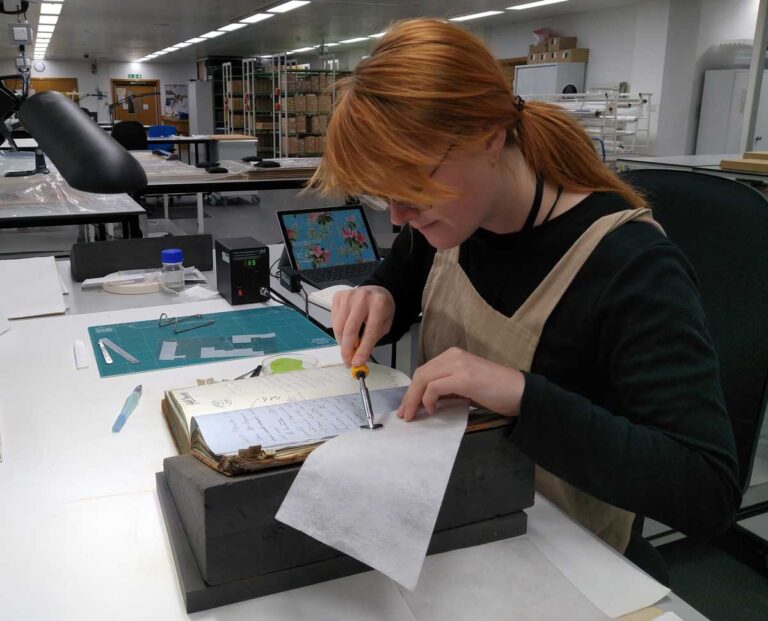A woman with red hair holds a small hand tool to a piece of tissue beside a large book.