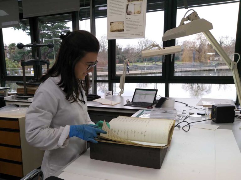 A woman in a white lab coat and blue gloves turns the page on a historical book, below two large table lamps.