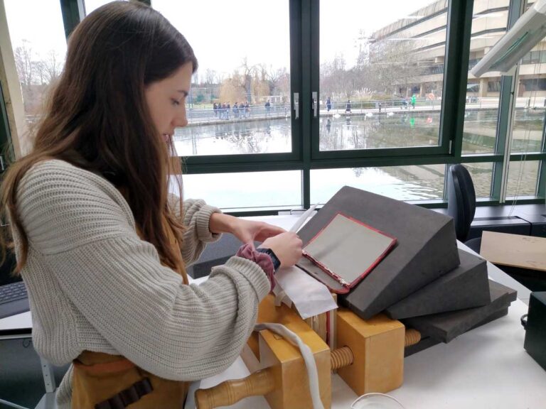 A woman works on the spine of a book, grey with a red lining, sitting on top of a pile of styrofoam blocks.