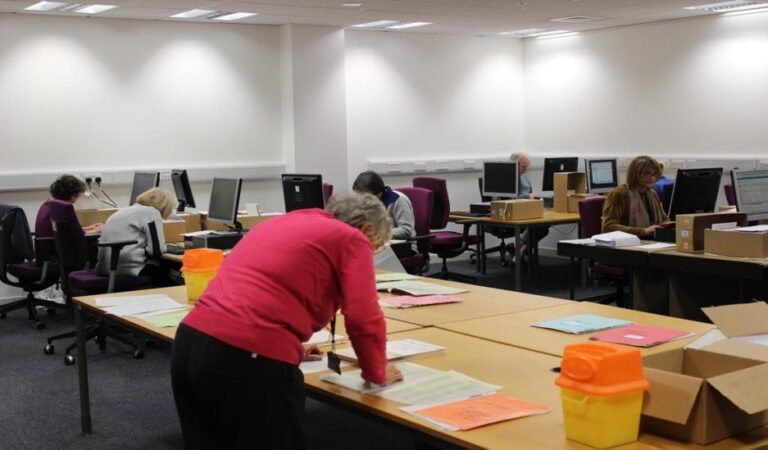 A group of volunteers working on a cataloguing project. A women in the foreground is reading documents on a table. Several people in the background work on computers