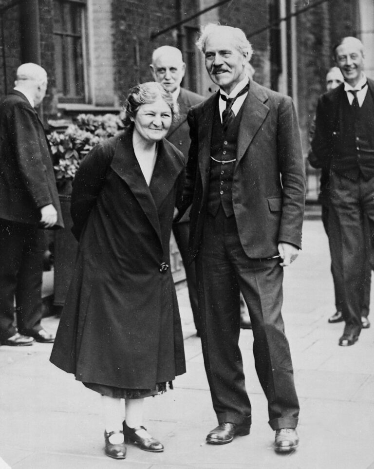 A black and white photograph of a man and woman standing next to each other smiling. There are four men in the background.