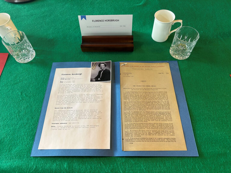 A file laid out on a green table with documents on Florence Horsburgh. There is an a picture of a woman and two glasses and a mug sit next to the file.