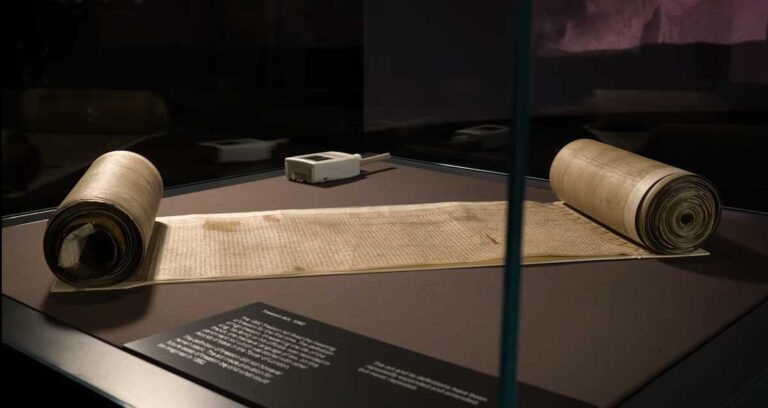 A manuscript, rolled up at either end, in a display case next to a small, white monitoring device.
