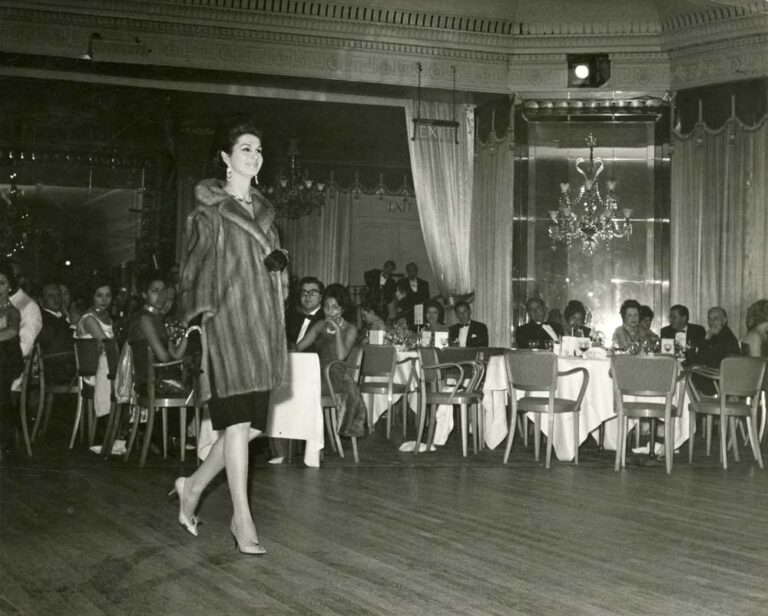 A woman in a fur coat walking next to tables with smartly-dressed people sat at them.