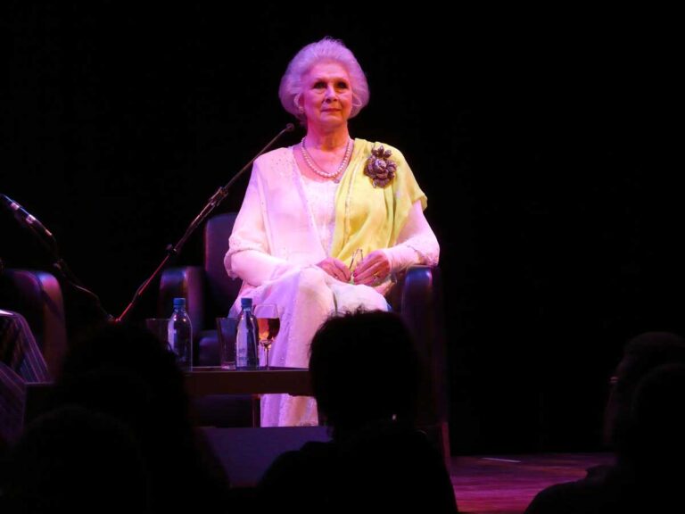 A woman with white hair, wearing a pearl necklace, sits in front of a microphone and an audience.