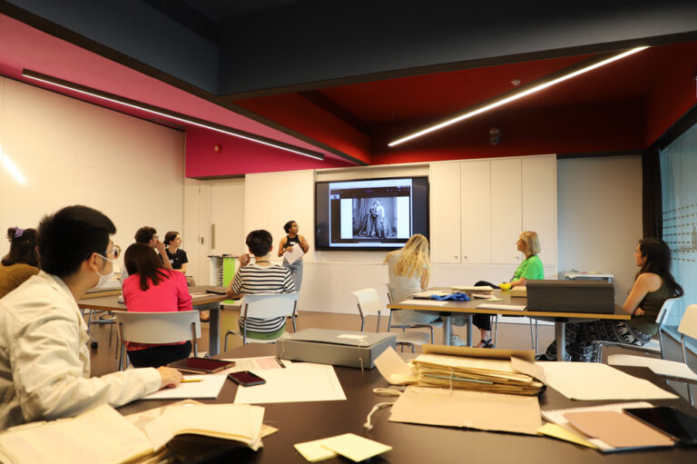 A group of students seated at tables in a classroom. One person presents in front of a screen. 