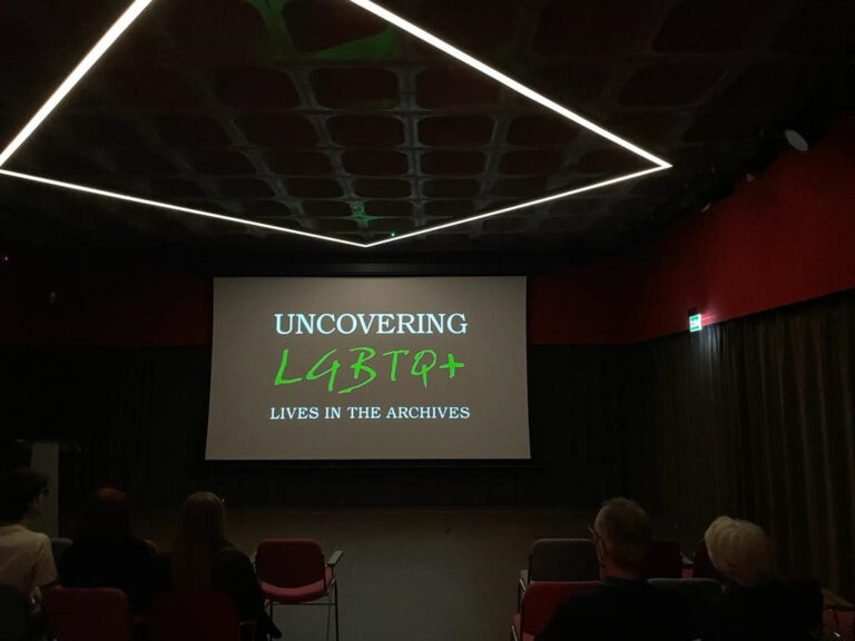 People seated in a cinema auditorium. On the screen are the words. 'Uncovering LGBTQ+ live in the archives'.