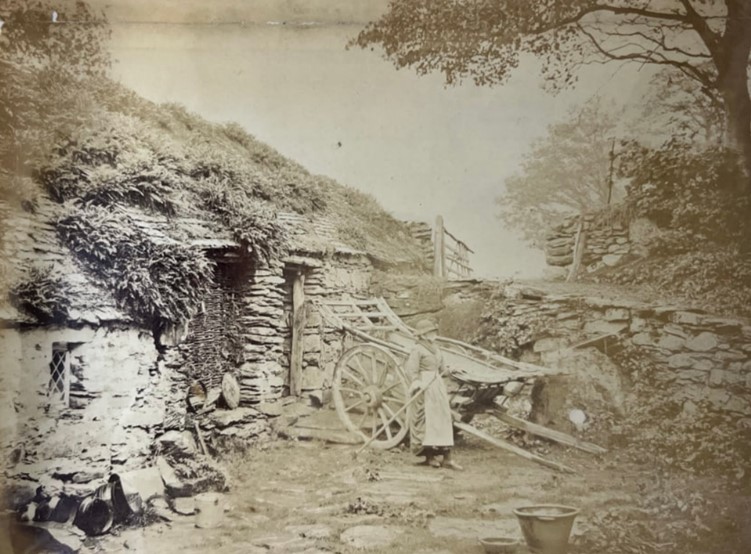 A black and white photograph of woman standing in front of a cart and a house in a rural setting.