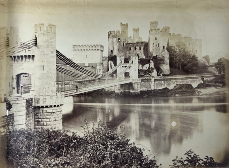 A black and white photograph of a castle across a bridge with a river running below. There is a man on the bridge. 