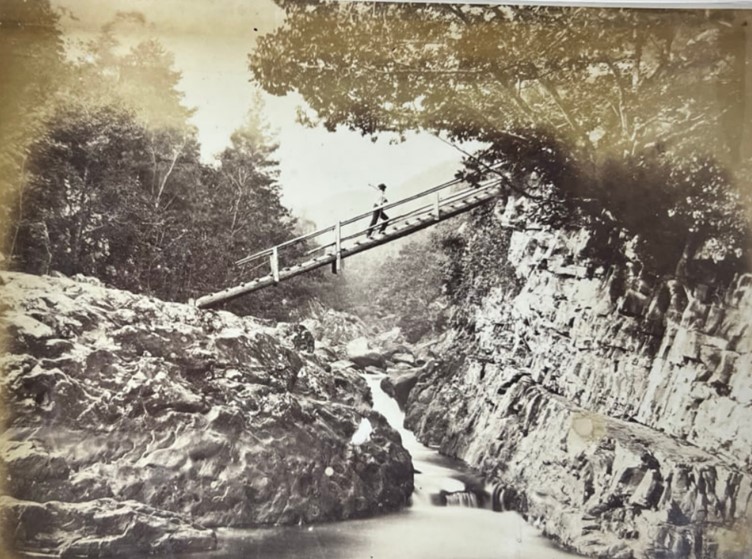 A black and white photograph of a man walking across a bridge which runs over a river, with rocks either side.