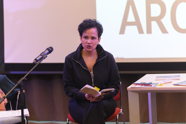 A seated woman reads from a book into a microphone.